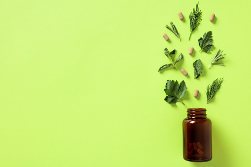 Amber glass bottle with vitamin pills in capsules and greenery on green background. Concept of healthy lifestyle, taking vitamins, nutritional supplements, minerals