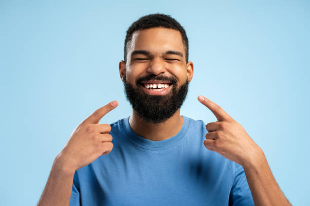 istock Happy African American man pointing with hands at teeth while closing eyes over blue background 2149593380