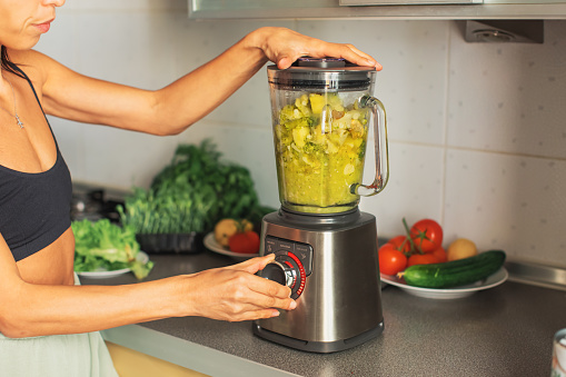 Young beautiful hispanic woman preparing vegetable smoothie pouring water on blender at the kitchen