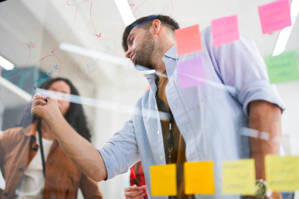 istock Business team uses a glass wall covered with sticky notes to outline processes during meeting. Engaged colleagues gather around a glass brainstorming board. plotting ideas and strategies. 2149597359