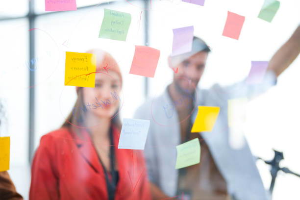 istock Business team uses a glass wall covered with sticky notes to outline processes during meeting. Engaged colleagues gather around a glass brainstorming board. plotting ideas and strategies. 2149597475