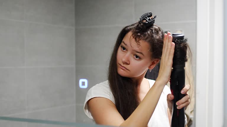Young woman winds curls on a brush attachment using a multi-styler hairdryer at home bathroom. The concept of hair care, fashion and beauty. Reflected view of the Mirror
