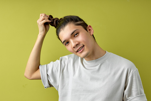 pleasant male model touching hair in topknot and looking at camera isolated on green, good-looking guy in casual light t-shirt smiling, having dark hair and white skin, hispanic or caucasian