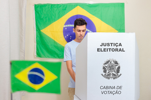 Brazilian man choosing his candidate on the electronic voting machine. On the wall, the Brazilian flag decorates the polling place. The image depicts the Brazilian elections