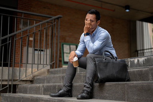 A stressed Asian millennial businessman, appearing tired and unhappy, sits on the steps outside a building, holding a coffee cup and covering his face, feelings of failure and exhaustion.