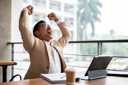 A cheerful Asian businessman in a beige suit and glasses raises his arms triumphantly while sitting at a table in a building corridor, celebrating success or good news received on his digital tablet.