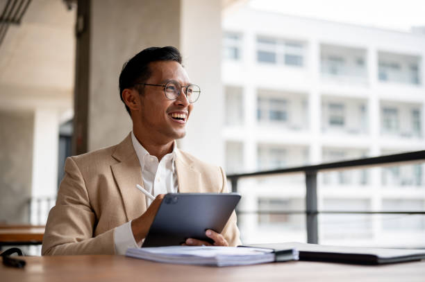 istock An optimistic Asian businessman smiles and looks away from the camera while holding a digital tablet 2149606821