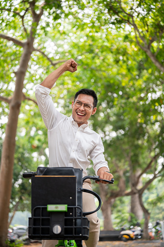 An excited Asian millennial businessman rides a bike triumphantly, raising his fist in celebration, having accomplished a goal or received good news, expresses joy while riding in a public park.