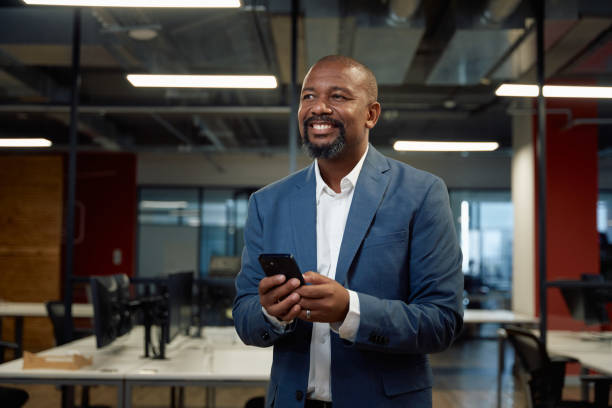 istock Mature black man in businesswear smiling while using mobile phone in office 2149611114