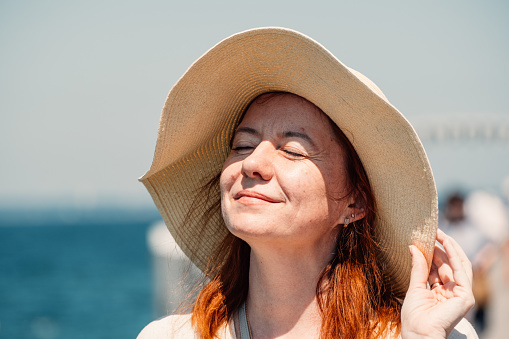 A red-haired woman enjoys a serene moment, eyes closed, with the sun on her face, under the shade of an elegant sun hat, epitomizing peaceful summer days