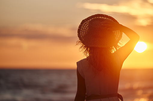 Rear view of a woman with sun hat standing on the beach and looking at sunset above the sea. Copy space.