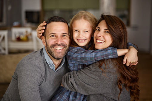 Portrait of happy parents and their small daughter embracing at home and looking at camera.