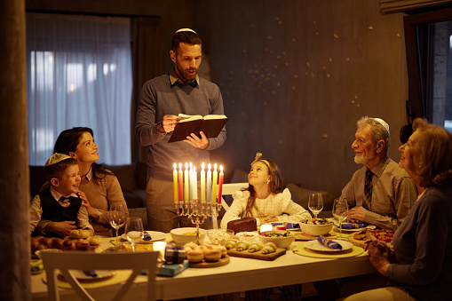 Mid adult man reading Torah before Hanukkah dinner with his extended family in dining room.