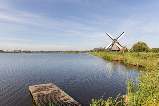 A classic windmill next to a lake under a bright, blue sky. A wooden jetty guides the viewer to the windmill, set amongst vibrant foliage. The still water and open setting create a peaceful, scenic view.