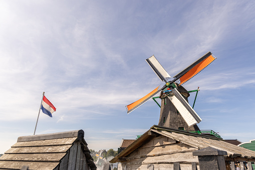 A windmill's striking silhouette against a soft blue sky, its blades adorned with the national colors. A proud Dutch flag flutters in the foreground, symbolizing national pride. The scene is framed with rustic wooden structures, possibly part of a mill complex, adding to the authentic atmosphere.