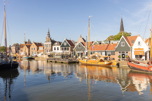 A Dutch canal scene, featuring historic architecture, quaint houses with gabled roofs, and a classic sailboat moored along the water's edge. A terrace with seating provides a welcoming spot for leisurely waterfront dining. The reflection of the buildings in the still water and the clear sky adds to the tranquil atmosphere.