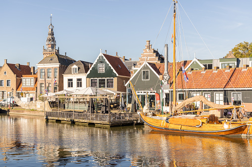 A Dutch canal scene, featuring historic architecture, quaint houses with gabled roofs, and a classic sailboat moored along the water's edge. A terrace with seating provides a welcoming spot for leisurely waterfront dining. The reflection of the buildings in the still water and the clear sky adds to the tranquil atmosphere