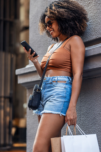 Shot of happy woman using smartphone while carrying shopping bags leaning against the wall in the city