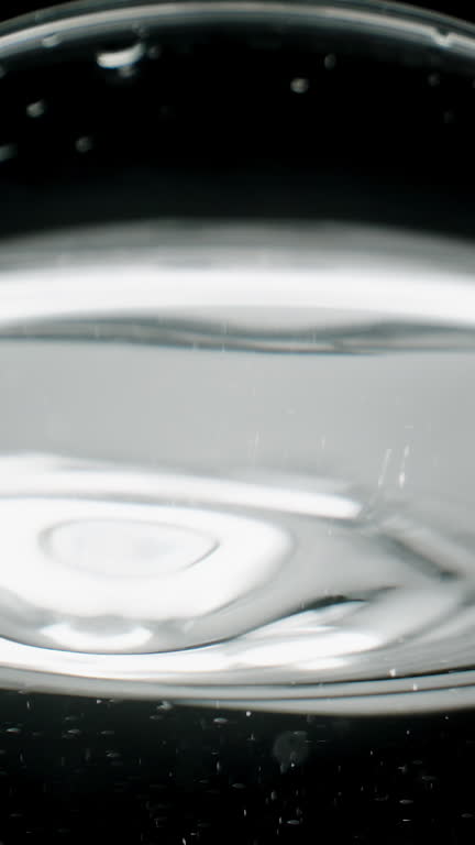 Vertical video. Water droplets dripping into the glass, macro view from a low angle on the silvery surface of the water resembling mercury.