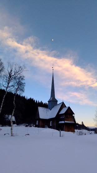 Winter view of Kvikne Kirke, Norway in the snow at sunset. The moon above the spire.