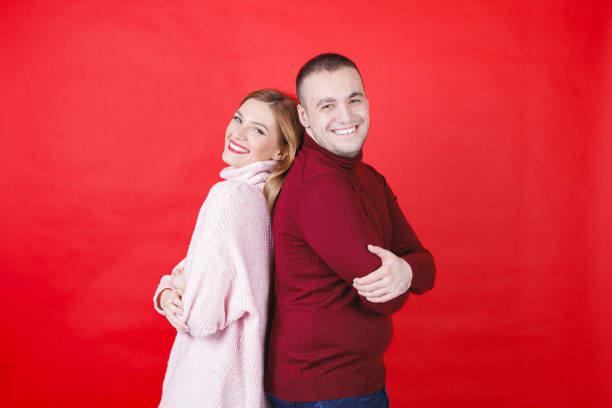 Young couple in turtleneck sweaters standing back to back and smiling at camera with joy, red background, studio shot