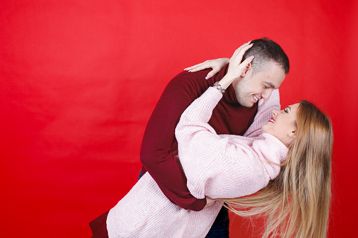 Young heterosexual couple in turtleneck sweaters hugging and leaning for old movie kiss, red background, studio shot
