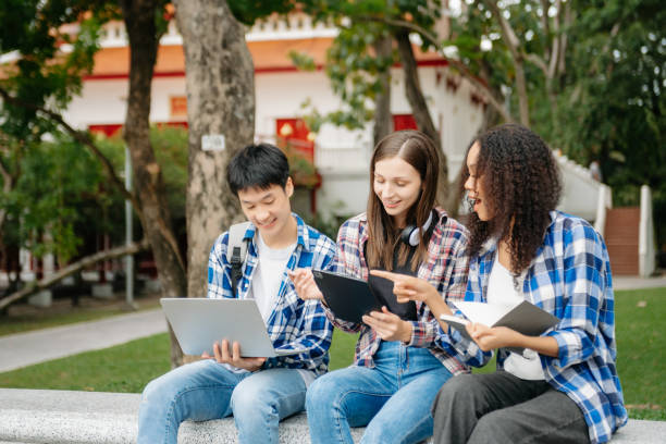 istock Three young college students is reading a book while relaxing sitting on grass in a campus park with her friends. 2149623273