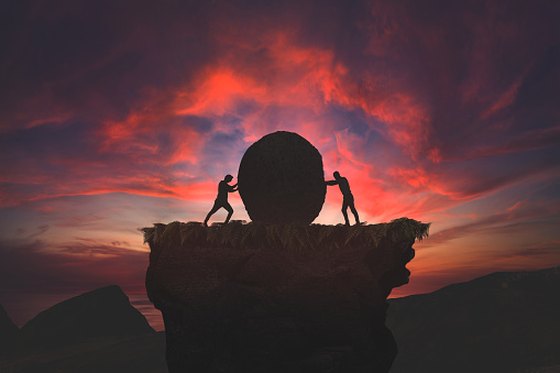 Two silhouetted figures push against a massive boulder atop a cliff, with a vivid sunset backdrop.