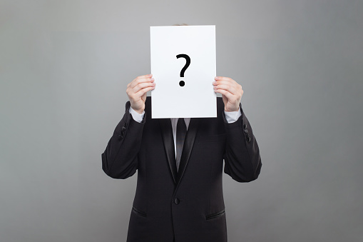 Business man wearing black suit hiding his face behind white paper with black question mark on grey background