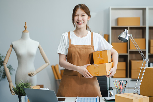 Young Asian SMEs woman holding parcels and looking to camera, standing among several boxes in modern office.