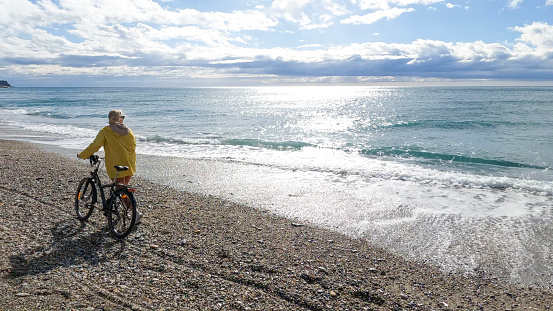 Mature woman pushes bicycle along empty beach, Liguria