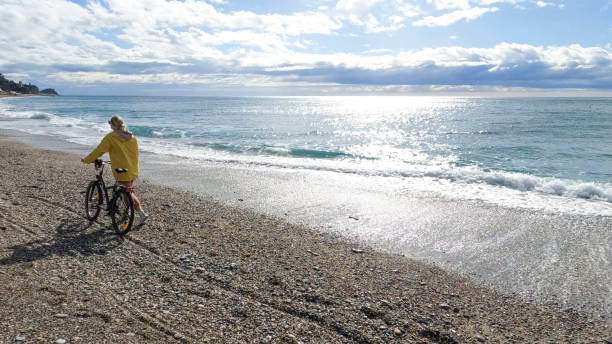 Mature woman pushes bicycle along empty beach, Liguria
