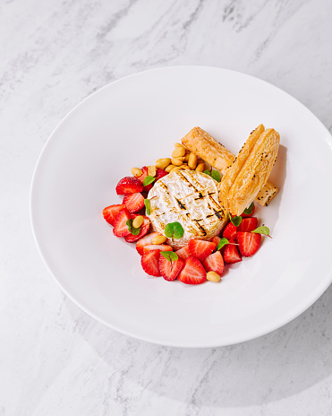 Exquisite and elegant gourmet bbq cheese presentation with plated sweet puff pastry and fresh strawberries on a white plate. Showcased in a top view on a marble background