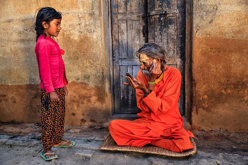 Little girl observing how Sadhu doing doing his makeup. In Hinduism, sadhu, or shadhu is a common term for a mystic, an ascetic, practitioner of yoga (yogi) and/or wandering monks. The sadhu is solely dedicated to achieving the fourth and final Hindu goal of life, moksha (liberation), through meditation and contemplation of Brahman. Sadhus often wear ochre-colored clothing, symbolizing renunciation.