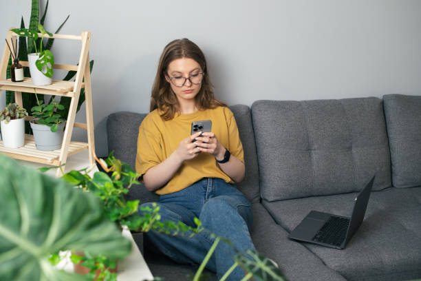 istock Girl talking on the phone while sitting on the sofa surrounded by indoor plants. Beautiful girl relaxing at home and looking at her phone. Jungle concept at home, remote work, freelancing 2149634043