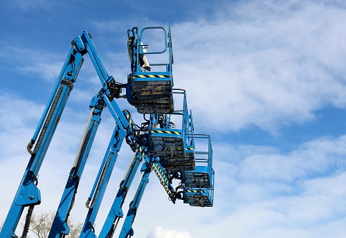 Five blue aerial work platforms of cherry pickers in a row against blue sky with clouds.