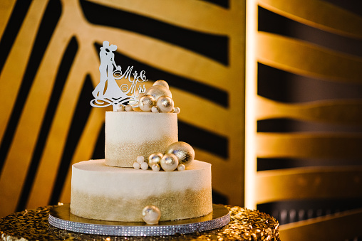 Wedding white cake decorated golden decor, figurines, topper bride and groom suite for a banquet. Selective focus. Delicious reception for luxury wedding ceremony. Trendy cake. Closeup. Mr and Mrs.