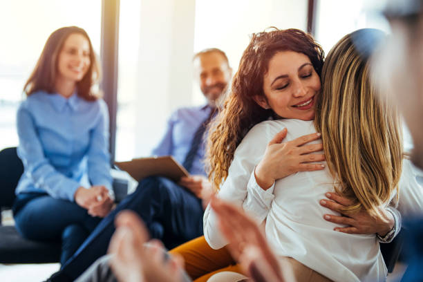 Support group patients hug each other during the therapy session The two women are attending a group therapy session. They are showing support and kindness. Portrait of female psychologist embracing young woman during therapy session in support group mercy hug stock pictures, royalty-free photos & images