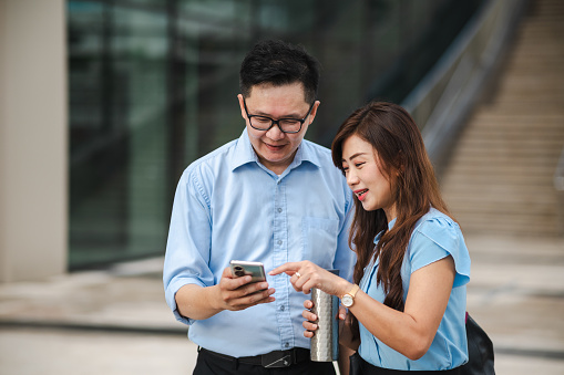 An Asian businessman and businesswoman standing outside an office building, engaged in small talk while using their phones.