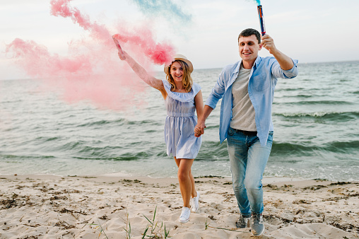 Man and woman having fun running on sand and holding blue and red, pink color smoke bombs and looking at camera on beach. Happy married couple holds hands smoke bomb. Baby shower. Twins:  boy and girl