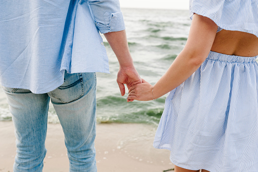Couple in love holding hands on seashore. Man and woman standing on sand. Back view. Female and male walk on beach ocean and enjoying a sunny summer day on vacation. Bottom down view on legs. Closeup