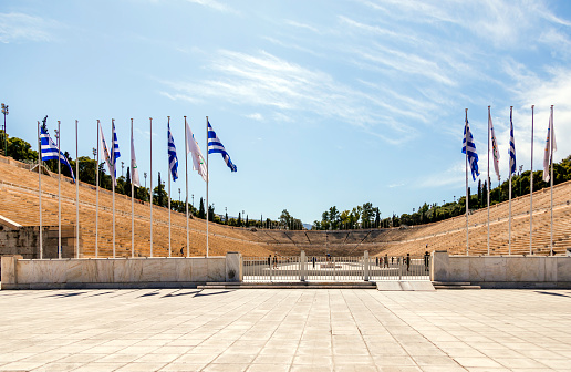 Pride of Greece: The Flag at Panathenaic Stadium. Beneath the radiant Athenian sun, the Greek flag proudly waves at the historic Panathenaic Stadium, also known as Kallimarmaro, in Athens, Attica. This iconic stadium, crafted entirely of gleaming marble, stands as a testament to Greece's enduring legacy. As visitors bask in the warm glow of a sunny summer day, they marvel at the grandeur of this multi-purpose arena, steeped in history and tradition. From hosting ancient athletic contests to modern Olympic events, the Panathenaic Stadium remains a symbol of Greece's enduring spirit and athletic prowess, drawing visitors from around the world to witness its timeless beauty and significance.