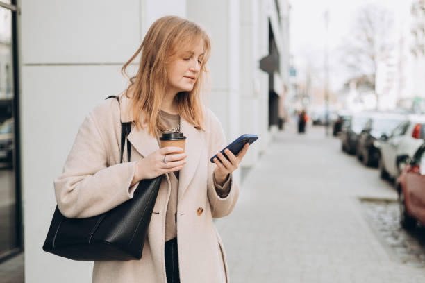 istock Beautiful smiling female with coffee cup using smartphone and standing outdoors. Phone Communication. Happy cheerful young woman walking on city street, Urban lifestyle concept. Traveler 2149648281