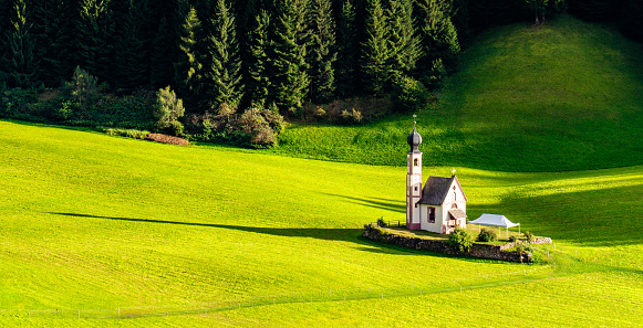 A small Church of St. John surrounded by lush green fields and backed by a dense forest. The bright sunlight casts natural hues on the scene, emphasizing the architectural and natural elements. Dolomites, Italy.