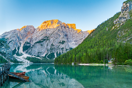 Pragser Wildsee features a serene view of crystal clear waters reflecting the majestic snow-capped mountains in sunrise rays, surrounded by a dense green forest; a wooden dock with boats.