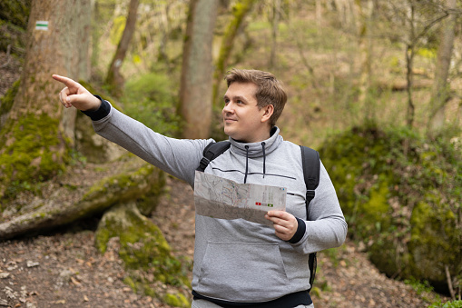 Happy men with paper map walking in forest. Blonde male in sport clothes pointing finger outdoors, he hiking on woodland