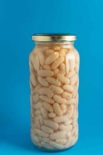 A clear jar filled with cooked white beans sits against a vibrant blue background. The beans are neatly packed, showcasing their smooth texture and light color