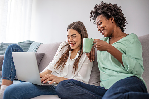 Two diverse friends relaxing on comfortable couch and watching film online on laptop device using home wireless internet.Multicultural young women working together on common blog at computer