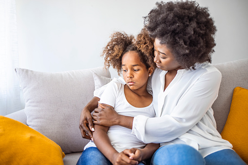 Black African mother embrace little preschool frustrated kid sitting on couch together at home. American loving mother supports disappointed daughter sympathizing, making peace after scolding concept