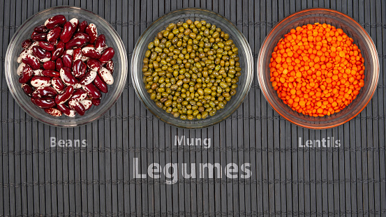 Three clear glass bowls are neatly arranged on a dark bamboo mat surface, each containing a different type of legume - red and white beans, green mung beans, and orange lentils.